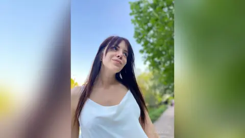 Young woman in a white sleeveless top smiling outdoors against a lush green summer background.