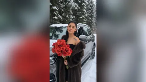 Elegant woman in a brown fur coat holding red roses by a luxury car in a snowy winter forest setting.