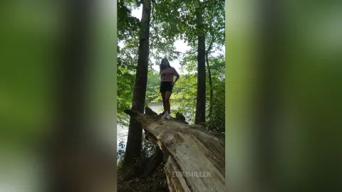 Young woman in casual summer outfit standing on a fallen log in a lush, sunlit forest setting.