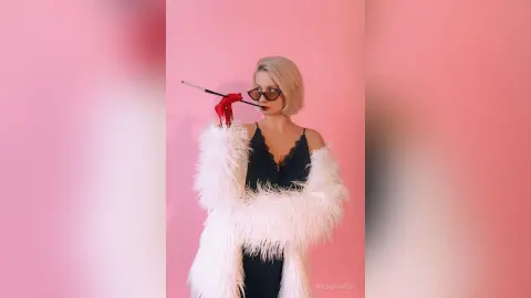 Glamorous blonde woman in a black lace dress and white faux fur coat posing against a pink background.