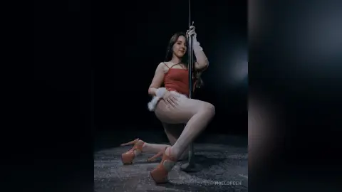 Woman in red bodysuit and white stockings posing gracefully by a pole in a dark studio setting.