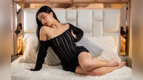 Young woman in a black knit off-the-shoulder mini dress relaxing on a white bed in a cozy bedroom setting.