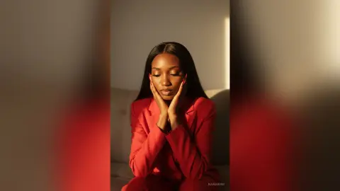 Elegant woman in a red blazer posing thoughtfully in warm, moody indoor lighting.