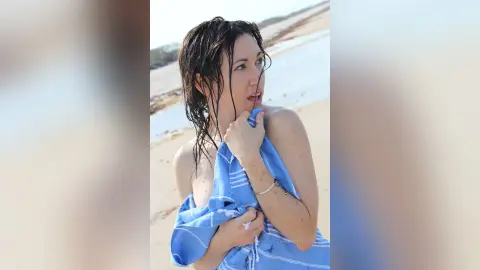 Woman wrapped in a blue striped towel on a sunny beach with wet hair and a serene mood.