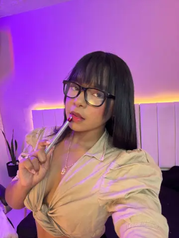 Young woman with bangs and glasses wearing a beige blouse in a room with purple mood lighting.