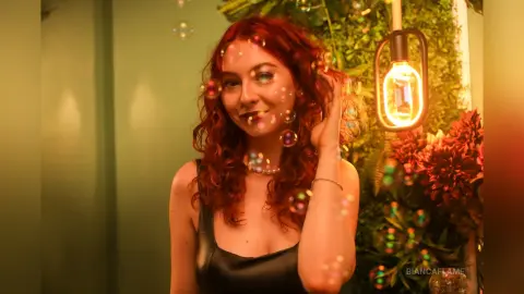 Smiling redhead woman in a black tank top surrounded by floating bubbles in a warm, moody indoor setting.