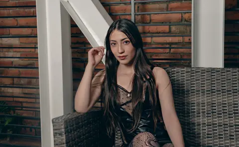 Young woman in a black lace outfit sitting on a wicker chair against a rustic brick wall background.