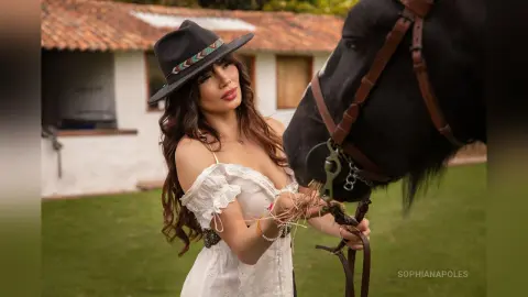 SophiaNapoles wears a black hat and white off-the-shoulder top while standing next to a black horse in a grassy outdoor setting.