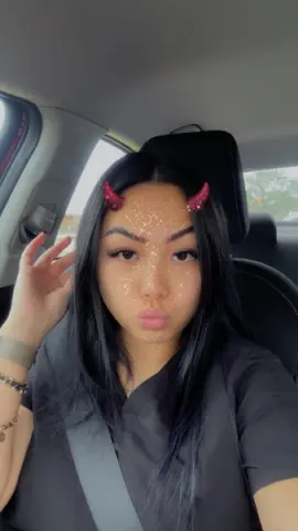 Young woman with glittery face makeup and red devil horns wearing a black shirt in a car.