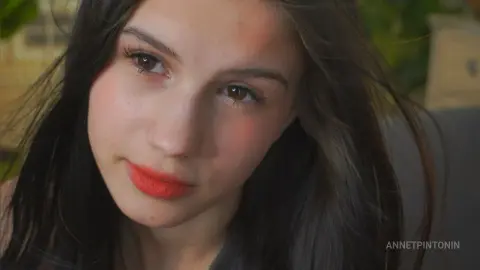 Close-up portrait of a young woman with long dark hair and red lipstick in a soft outdoor setting.