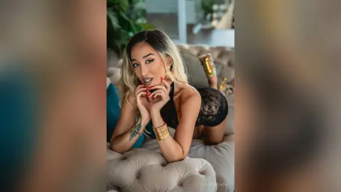 Glamorous woman in black lace bodysuit and gold jewelry relaxing on a tufted sofa in a bright room.