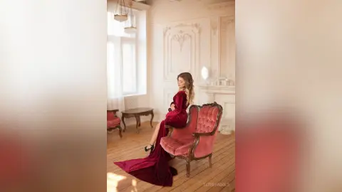 Elegant woman in a long red velvet gown sitting in a vintage pink armchair within a bright, classical room.