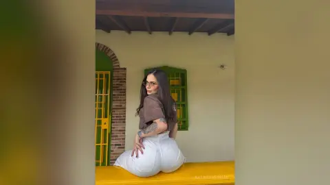 Young woman with tattoos in a brown t-shirt and white pants sitting outdoors by a vintage green door.