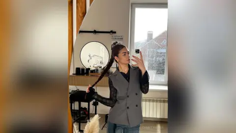 CharityRobes takes a mirror selfie in a grey blazer and black mesh sleeves, posing with her long dark braid in a bright room.