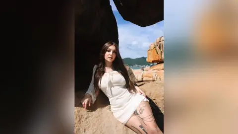 Young woman in a white dress sitting on rocks by the ocean under a bright blue sky.