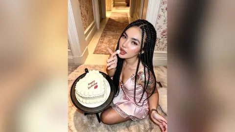 Smiling woman with long braids in a pink satin slip dress holding a happy birthday cake in a cozy room.