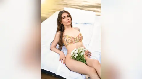 Young woman in a floral lace bikini relaxing on a white floating mattress in a serene lake setting.