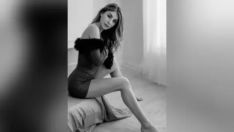 Elegant woman in a black off-the-shoulder dress sitting on a bed in a bright, minimalist room.