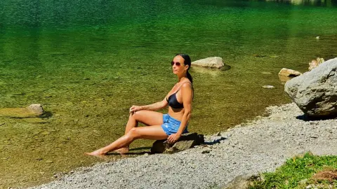 Woman in black bikini top and denim shorts relaxing on a rocky shore by a clear turquoise mountain lake.