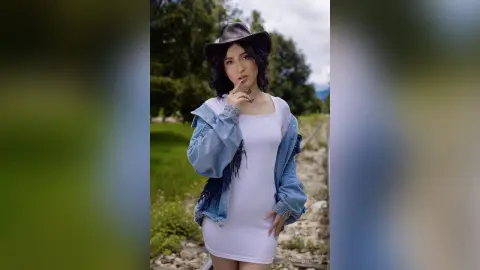 Young woman in a white dress and denim jacket wearing a black cowboy hat in a lush green outdoor setting.