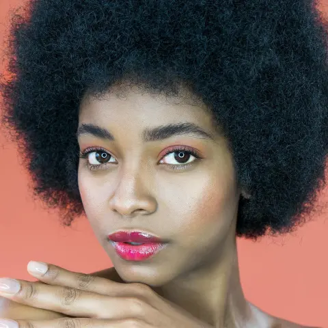 Beautiful Black woman with natural afro hair and glowing makeup posing against a warm peach background.
