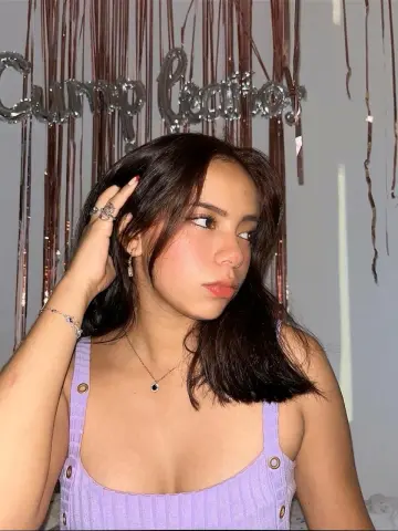 MilaRossee poses in a purple tank top with her hand in her long dark hair against a silver fringe background.