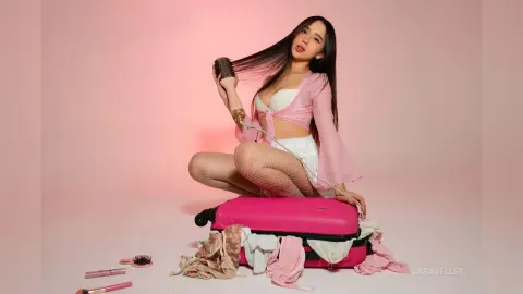 Young woman in a pink crop top and white skirt sitting on a pink suitcase against a soft pink background.