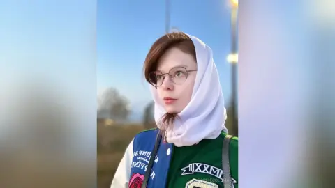 Young woman in a white headscarf and green graphic jacket looking thoughtfully at a sunset beach landscape.