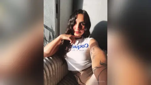 RossiNoleen wears a white tank top and tan shorts while sitting against a wall with her long dark hair falling over her shoulder.