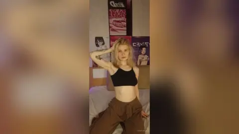 Blonde woman in a black crop top and brown trousers posing in a cozy bedroom with anime posters.