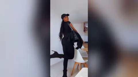 Stylish woman in a black outfit and cap posing confidently in a bright, modern minimalist room.