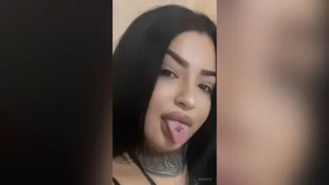 Close-up portrait of a young woman with long dark hair and neck tattoos making a playful expression indoors.