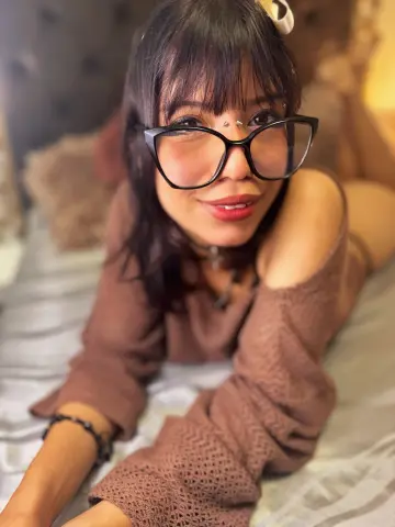 Smiling woman with black bangs and glasses wearing a brown knit sweater relaxing on a white bed.