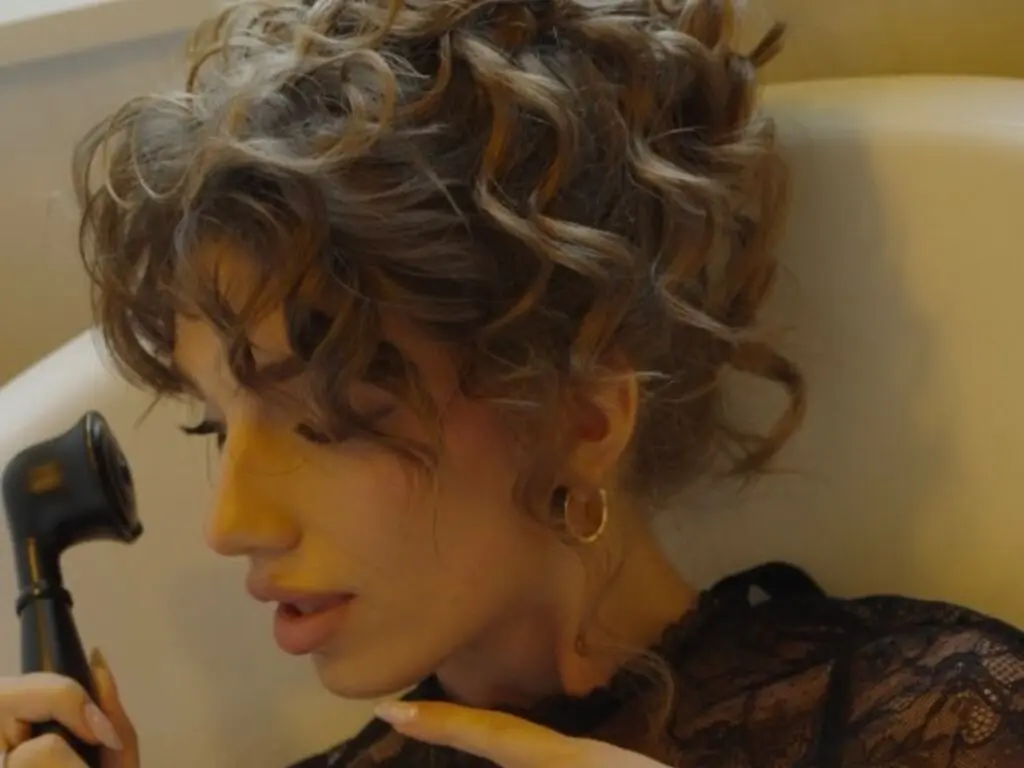 Woman with curly brown hair styling her hair in a bathroom with gold hoop earrings and a black lace top.
