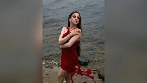 Elegant woman in a flowing red dress posing on a moody, rocky beach by the ocean.