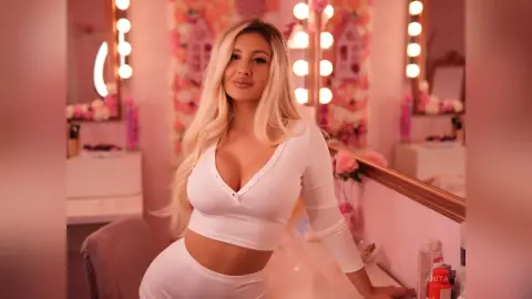 Anita poses in a white long-sleeved crop top in front of a lit vanity mirror.