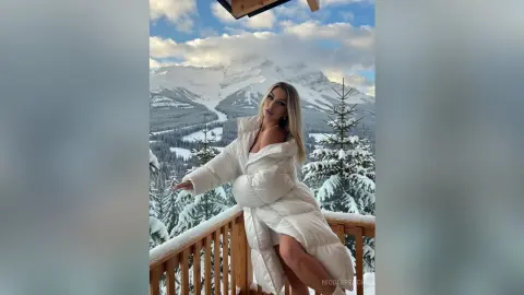 NicolePeach poses on a snowy wooden balcony wearing a white puffer coat that shows off her long legs against a mountain landscape.