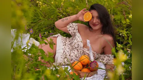 Smiling woman in a floral sundress relaxing in a lush green meadow with a basket of fresh oranges.