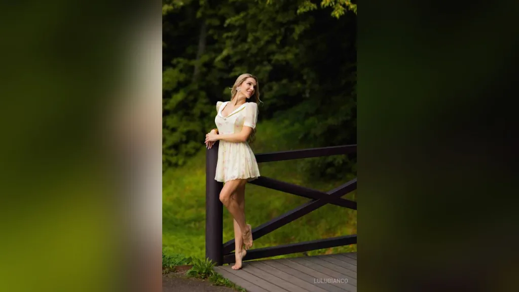 LuluBianco poses in a white feathered outfit and sheer black gloves with her long blonde hair flowing over her shoulder.