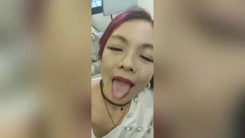 OishiMeily — Playful woman with pink hair in a white top sticking her tongue out in a cozy indoor setting.