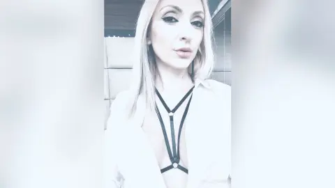 Blonde woman in a white shirt and black harness necklace posing in a bright, modern indoor setting.