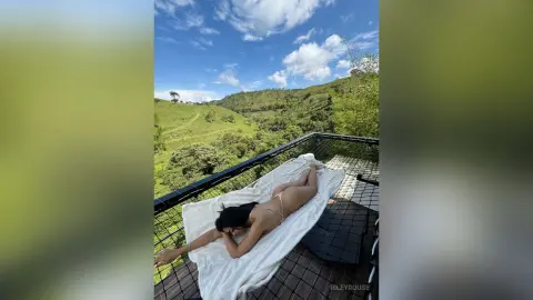 Relaxed woman in a bikini sunbathing on a white lounge chair overlooking a lush green mountain valley.