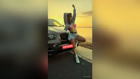 Cheerful woman in denim shorts and blue shirt posing by a car during a golden hour sunset.