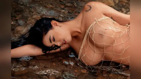 Nude woman in white fishnet mesh outfit lying on dark river rocks in a moody outdoor setting.