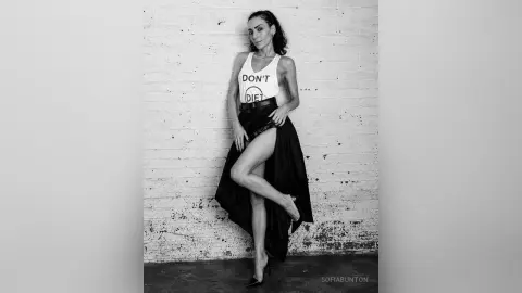 Stylish woman in a white graphic tank top and long black skirt posing against a white brick wall.