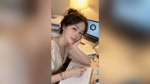 Young woman in a white lace blouse studying at a desk with a laptop and notebook in a cozy, warm room.