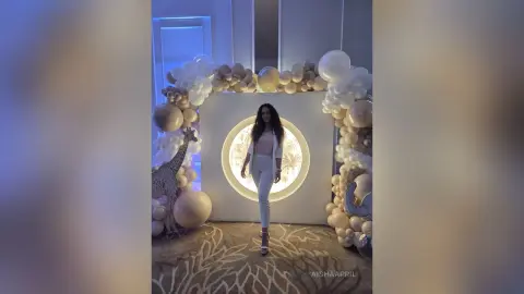 AishaApril stands in front of a large circular light, wearing a white top and white pants, surrounded by white and tan balloons.