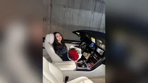 Young woman in a striped shirt sitting in a luxury car interior holding a bouquet of red roses.