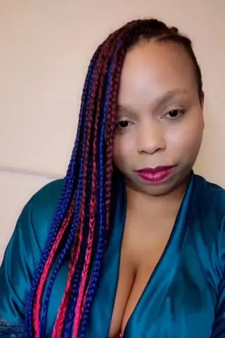 Woman with long blue and red ombre braids wearing a teal satin blouse in a bright indoor setting.