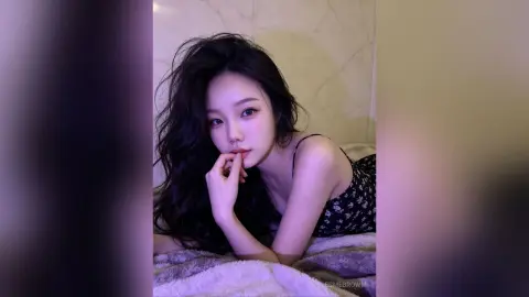 Young woman with long wavy hair in a black floral camisole relaxing on a cozy bed in soft purple lighting.
